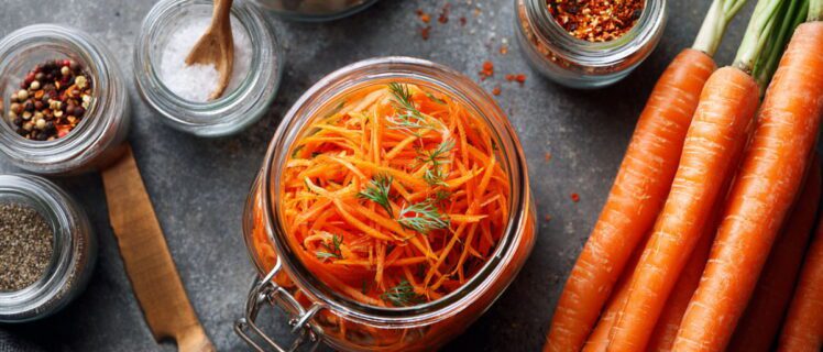 Салат моркви по-корейськи в банці, поруч цілі моркви та баночки зі спеціями